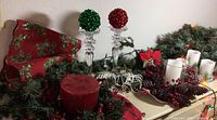 Full display of holiday garlands, white candles, floral centerpiece with red berries and pine cones, large red pillar candle, and two tall clear glass candlesticks holding red and green shiny ornament balls.