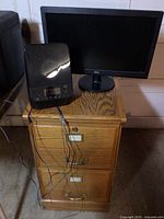 Wood file cabinet with three drawers, radio/CD player on top, and two computer monitors behind it