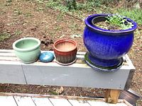 Four ceramic pots arranged on a ledge: large blue glazed pot with saucer, medium green pot, small brown and red pot with saucer, plus a small turquoise glazed pot and a small blue decorative rock