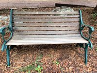 Front view of a weathered wooden slat park bench with a green metal frame and armrests, situated outdoors on natural ground with leaves and twigs.