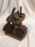 Five smoking pipes arranged on and around a two-tier wooden stand with circular pipe holes, showing the shape and condition including one bent pipe stem.