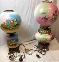 Both antique hurricane lamps shown side by side with cords attached, decorative hand-painted glass globes and ornate metal bases in view.