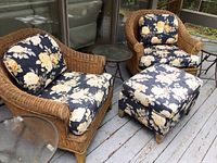 Two wicker armchairs with floral cushions, ottoman, and one small round metal and glass table shown on wooden deck.