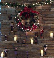 Mantle setup with 8 wooden nutcrackers of varying sizes, lighted wreath with red bow and decorations, and lighted garland with white lights.