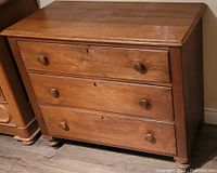 Front and angled view of antique three drawer dresser showing natural wood finish, round knobs, keyholes, and solid wood construction.