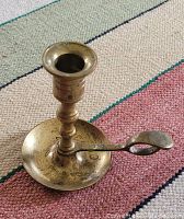 Photo showing a small brass candle holder with a finger loop handle, placed on a textured fabric surface.