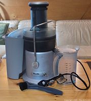 Full view of Breville juicer with attached pulp container, clear juice jug with handle, power cord, and cleaning brush on wooden surface with beige sofa in background.