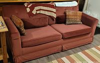 Burgundy fabric sofa bed seating three, soft fabric upholstery, two large seat cushions, and four assorted matching cushions. Sofa has a skirted base and button-tufted back with arms on both sides.