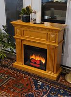 Oak finish electric fireplace with flame effect on a patterned rug, placed in front of a window, with decorative plants and a candle on top.