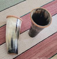 Two hollowed out horns used as drinking cups, placed on a striped textile background showing their shape, size, and decorative braided rims.