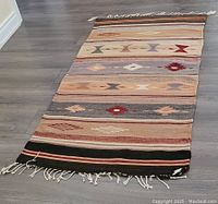 Photo of a short woven runner rug laid out on floor showing full length. Rug features multiple horizontal stripes with geometric and tribal-inspired motifs in earth tone colors and fringe tassels at both ends.