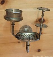 Brass wall mounted sink caddy showing soap dish, toothbrush holder, and cup holder attached to wooden background.