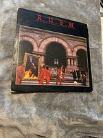 Front cover of Rush 1975 self-titled vinyl LP album showing artistic photo of people carrying paintings in front of a stone building with arched windows.