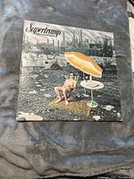 Front cover of Supertramp's Crisis? What Crisis? album featuring a man in sunglasses sitting under an orange and white umbrella in an urban industrial setting.