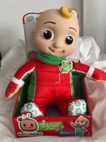 Front view of the Cocomelon JJ plush doll in red snow suit seated in original packaging.