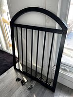 Black metal gate leaning against a door, showing vertical bars with an arched top, accompanied by small mounting hardware on the floor.