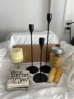 Photo showing three tall black metal candle holders, a clear glass candle holder with a cream pillar candle inside, two beige pillar candles, one jar candle with a metal lid, and two ceramic coasters with black text.