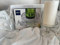 Overview photo of the Mikasa crystal wine server base accompanied by a stopper, candle on holder, and stemless wine glass placed against a white fabric background with product box behind.