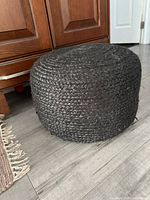 Photo showing front angle of black woven pouf footstool beside wood cabinets on gray floor.