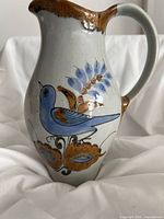 Front side view of a beige ceramic pitcher with brown rim and handle, featuring a hand-painted blue bird and floral motifs.