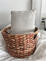 Front and side views of a round handwoven basket with natural and reddish fiber, containing a beige decorative pillow with sparkle.