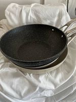 Photo showing one black and two cream-colored frying pans stacked, highlighting The Rock's speckled non-stick surface and silver handles.