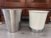 Two metal trash bins side by side, one stainless steel and one white-coated with stainless steel base.