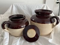 Two ceramic bean crocks with one lid placed in front, showing a brown glazed top half and beige bottom half, with one crock larger and one smaller both having handles.