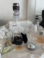 Clear acrylic Kuraidori folding wine decanter with two wine glasses and five perfume bottles of different shapes and fill levels, on white cloth.