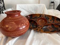 Photo of a round wooden platter with carved floral and abstract patterns alongside a terracotta-colored ceramic vase featuring etched fish motif.