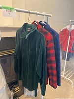Three hanging tops visible, green velour cardigan, red plaid blazer, and blue plaid blazer.