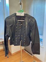 Front view of the black leather military-style cropped jacket with double rows of brass-tone snap buttons down the front. Several buttons are missing.
