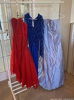 Three gowns on a rack: red strapless J. McClintock prom dress with beading, blue long-sleeve beaded gown, light lavender strapless satin gown
