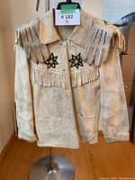 Front view of beige suede jacket with collar, fringe across chest and matching gloves draped over shoulders