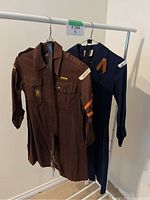 Image showing two vintage Brownie Girl Guide uniforms hanging on a rack: one brown and one dark navy, both with badges and patches.