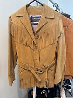 Front view of the vintage light brown suede jacket with fringe on chest and arms, button closure, and belt.