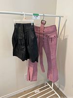 Pink suede pants with matching belt and black leather shorts hanging on a rack.