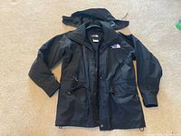 Full front view of the black women's Gore-Tex jacket by The North Face, laid flat showing front pockets, button and zip closure, and hood.
