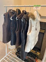 Five women's vests hanging on a rack: white fur-trimmed padded vest, several brown and black quilted and smooth fabric vests.