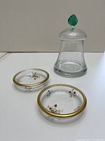 Clear glass bell-shaped candy dish with removable lid topped by green handle, alongside two small round glass plates with gold trim and floral designs.