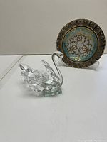 Clear faceted glass swan figurine with smooth neck and faceted wings displayed next to a small decorative metal and ceramic plate with blue and gold design.