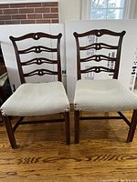 Two matching dark wood dining chairs with four horizontal slats and decorative cutout on backrest. Worn off-white upholstered seats, wooden legs with stretchers.