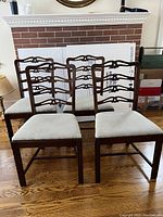 Four dark wood dining chairs arranged in two rows showing carved backs and light fabric upholstered seats against a wood floor and brick backdrop.