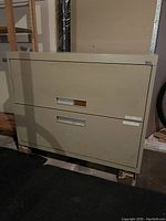 Photo shows beige 2-drawer metal lateral filing cabinet with recessed handles and labels, placed in a storage space with shelving and other items around.