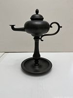 Full view of matte dark metal decorative lamp resembling an old teapot with curved handle and long spout, resting on a pedestal base.
