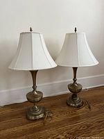 Pair of matching antique-gold colored table lamps with cream tulip-shaped fabric shades on a wooden floor against a white wall.