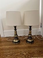 Pair of matching desk lamps with gold and black bases and taupe shades on a hardwood floor against a white wall.