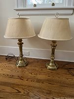 Two matching brass colored table lamps approximately 26 inches tall with beige fabric lampshades on hardwood floor.