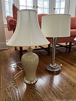 Both lamps shown side by side on a wooden floor with a couch in the background. The beige ginger jar lamp has a fabric tulip-shaped shade, and the chrome lamp has a sleek cylindrical metal body with a barrel shade.