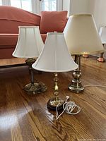 Three brass-colored desk lamps of varying sizes shown together on a hardwood floor with fabric shades.